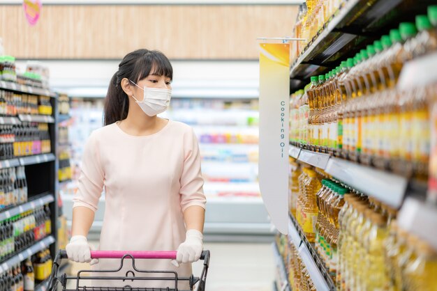 Aziatische vrouw met hygiënisch masker en rubberen handschoen met winkelwagen in kruidenier en op zoek naar bakfles om te kopen tijdens covid-19-uitbraak ter voorbereiding op een pandemische quarantaine