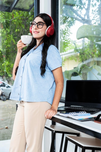 Aziatische vrouw met lenzenvloeistof luisteren naar muziek met koptelefoon en houden een kopje koffie met een laptop en een notebook met een mobiele telefoon op tafel