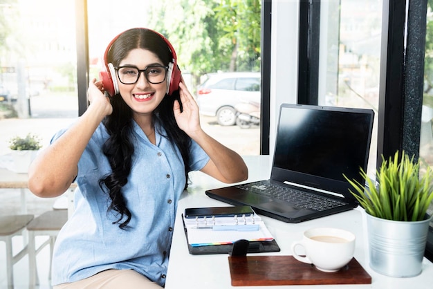 Aziatische vrouw met lenzenvloeistof luisteren naar muziek met koptelefoon met een laptop en een notebook met een mobiele telefoon en een kopje koffie op tafel