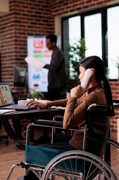 Aziatische vrouw met lichamelijke beperking die op een vaste telefoon praat, op afstand een gesprek voert op de kantoortelefoon. Rolstoelgebruiker met chronische gezondheidstoestand chatten op telefoongesprek.