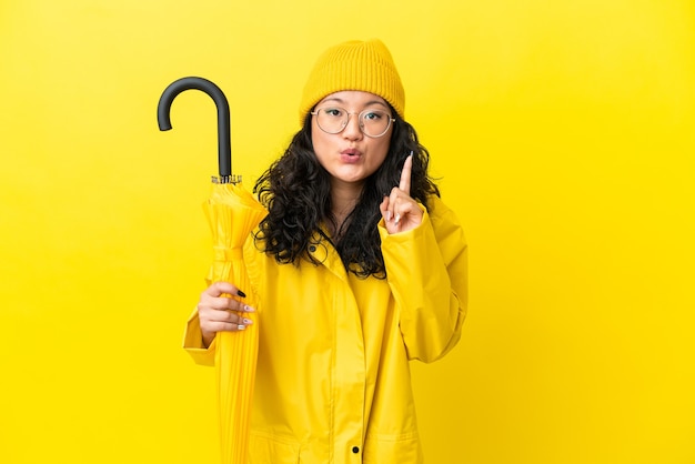 Aziatische vrouw met regenbestendige jas en paraplu geïsoleerd op een gele achtergrond die van plan is de oplossing te realiseren terwijl ze een vinger opheft