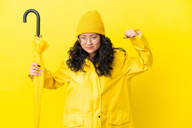 Aziatische vrouw met regendichte jas en paraplu geïsoleerd op gele achtergrond die sterk gebaar doet