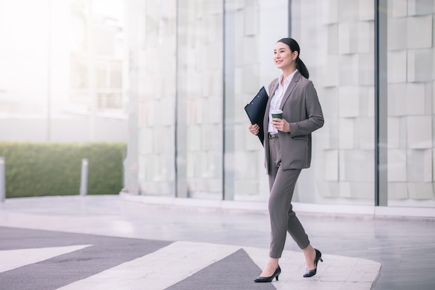 Aziatische vrouw met smartphone die zich tegen straat vaag gebouw bevindt. Mode zakelijke foto van mooi meisje in casual suite met telefoon en kopje koffie