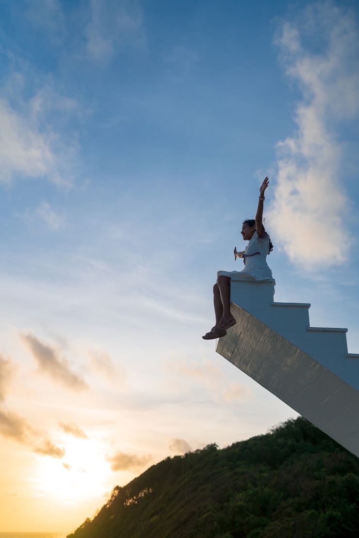 Foto aziatische vrouw steekt armen omhoog, zit bovenop een houten trap met avondrood op de achtergrond, overwinning, succesconcept