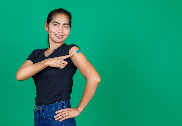 Aziatische vrouw van middelbare leeftijd die lacht en haar arm laat zien met een pleister die laat zien dat ze is ingeënt voor het Covid 19-virus op een groene achtergrond. Concept voor Covid 19-vaccinatie.