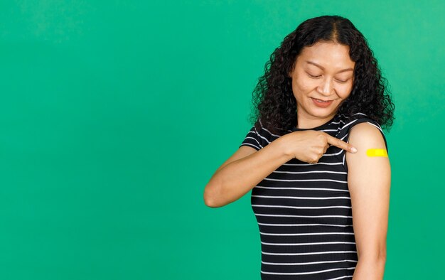 Aziatische vrouw van middelbare leeftijd die lacht en naar haar arm wijst met een pleister die laat zien dat ze is ingeënt voor het Covid 19-virus op een groene achtergrond. Concept voor Covid 19-vaccinatie.