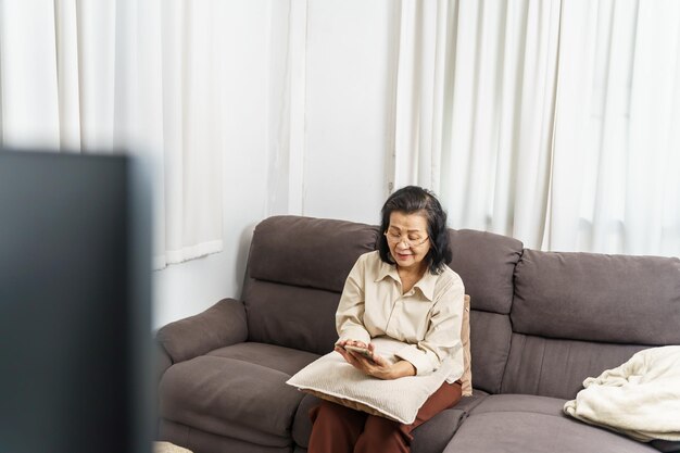 Aziatische vrouw van middelbare leeftijd met een mobiele telefoon, oudere vrouw met een slimme telefoon, bejaarde vrouw die thuis een smartphone gebruikt met een technologisch concept