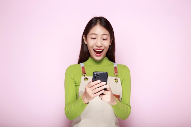 Aziatische vrouw ziet er verbaasd uit terwijl ze de telefoon in haar hand houdt Portret van een mooie jonge vrouw op een lichtroze achtergrond gelukkig en glimlachend in staande positie