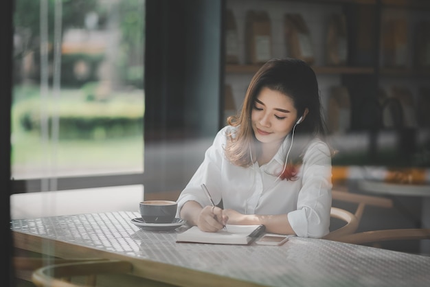 Aziatische vrouw zit en gebruikt een pen om op een papieren notitieboekje te schrijven met een laptopcomputer en draagt oortelefoons. Werk overal concept. Werk vanuit huis concept.