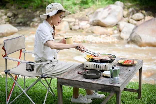 Aziatische vrouw zit in de buurt van de rivier en bereidt de lunch voor op het draagbare campingconcept van de picknickkachel