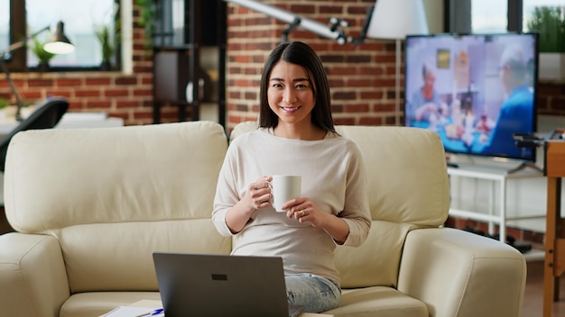 Aziatische vrouw zit in de woonkamer en werkt vanuit huis terwijl ze naar de camera glimlacht. Gelukkige student in modern appartement met laptop op tafel terwijl hij wacht tot de online les begint.