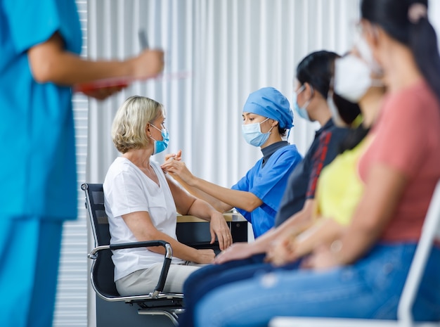 Aziatische vrouwelijke arts draagt een gezichtsmasker en ziekenhuisuniform die een coronavirusvaccin injecteert dat is geschoten op de schouder van een blanke oudere vrouw, terwijl andere multi-etnische patiënten in de wazige wachtrij op de voorgrond kijken.