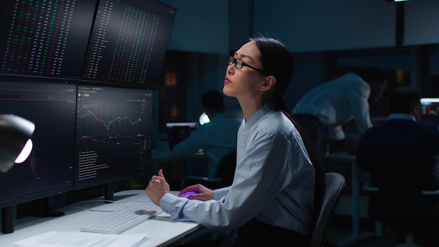 Aziatische vrouwelijke handelaar kijkt naar werkstation met meerdere schermen met grafieken en gegevens die laat in een donker kantoor werken