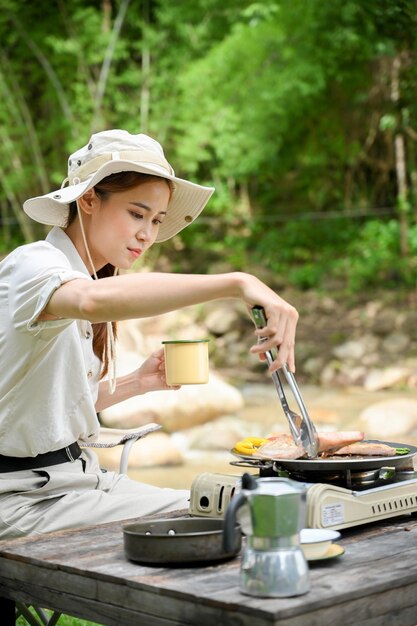 Aziatische vrouwelijke kampeerder die haar lunch kookt en een varkenskotelet op een picknickpan roostert