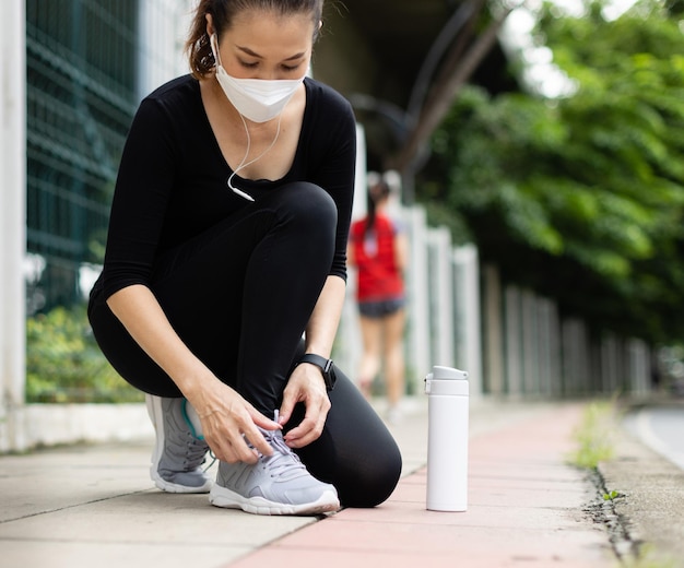 Aziatische vrouwelijke loper die een hygiënisch beschermend gezichtsmasker draagt en een schoentouw vastbindt tijdens een training in het stadspark. Concept van gezondheidszorg in de tijd van de uitbraak van het coronavirus.