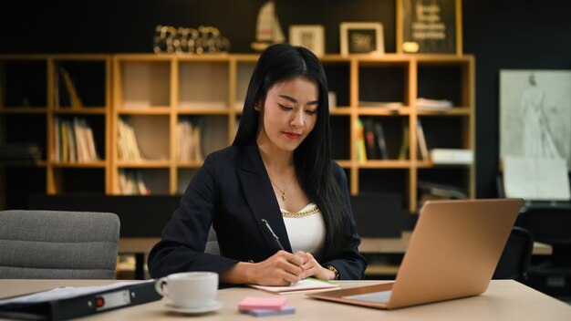 Aziatische vrouwelijke manager die met laptop werkt en haar businessplan op kladblok schrijft