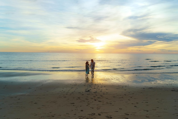 Aziatische vrouwelijke reiziger die de hand van de man vasthoudt en er een prachtige zonsondergang op het strand uitziet, koppel op vakantie in de zomer en vrijheidsconcept