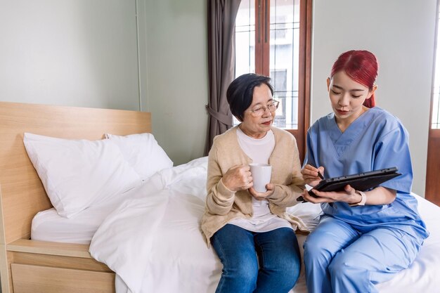 Aziatische vrouwelijke verpleegster die scrubs draagt, rapporteert de gezondheidsstatus van Senior Aziatische vrouw met digitale tablet