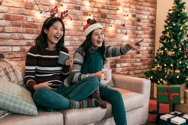 Aziatische vrouwelijke vrienden kijken naar film en hebben plezier op kerstavond zittend op de bank met geschenkdozen onder de kerstboom. jonge meisjes wijzen vrolijk naar een komedie op het televisiescherm waarop te zien is dat er wordt gelachen