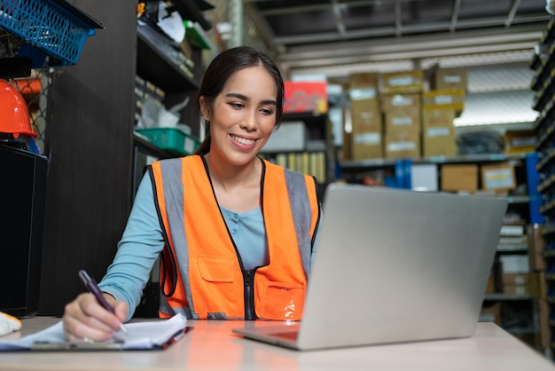 Aziatische vrouwelijke werknemer in veiligheidsvest zit en gebruikt een laptop die werkt op het kantoor van de magazijnfabriek