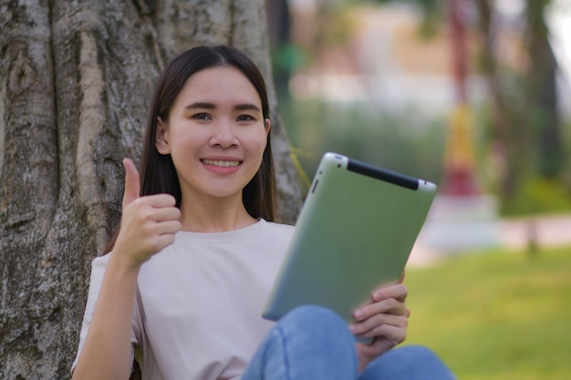 Aziatische Vrouwen die tablet op sociaal netwerk houden en online door Internet-zitting in stadspark winkelen