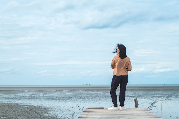 Aziatische vrouwenreiziger die zich bij zeegezicht op houten brug met blauwe hemel en oceaan bij achtergrond bevinden