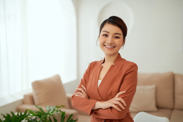 Aziatische werkende vrouw die een zwart pak en kruisarmen draagt, staat in een witte kantoorruimte,