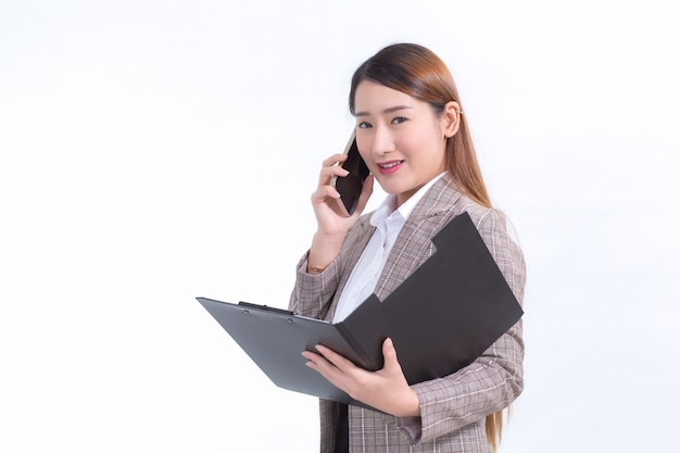 Aziatische werkende vrouw in formeel pak met wit overhemd belt telefoon en opent documentbestand of klembord om gegevens te controleren.