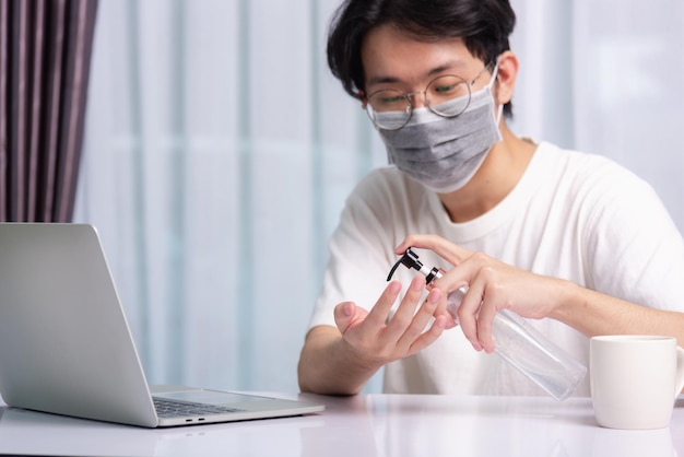 Aziatische zaken jonge knappe man bril werkend vanuit kantoor aan huis hij plaatst ziekte coronavirus of COVID-19 in quarantaine met beschermend masker en handen reinigen met ontsmettingsgel op laptop aan de voorkant