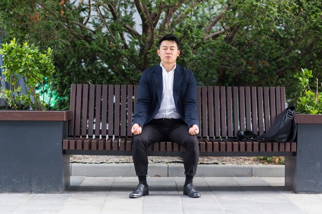 Aziatische zakenman die ademhalingsoefeningen uitvoert om stress te kalmeren, zittend op een bank tijdens een lunchpauze in een pak