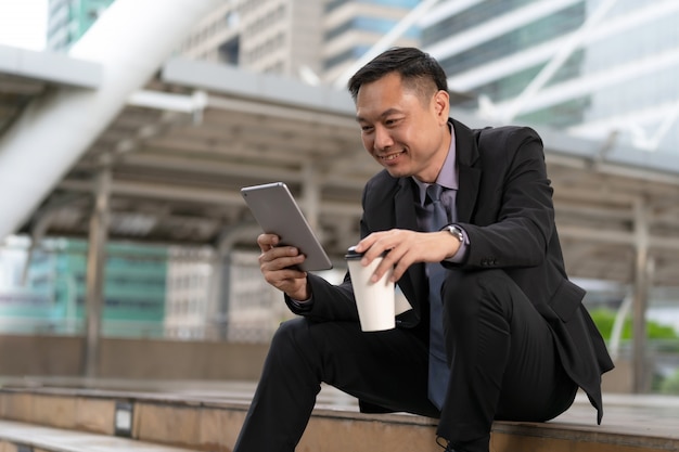 Aziatische Zakenman die en digitale tablet met bedrijfsbureaugebouwen zitten houden in de stad