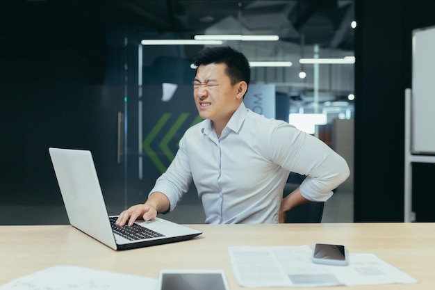 Aziatische zakenman die op kantoor werkt met ernstige rugpijn oververmoeide werknemer die met laptop werkt
