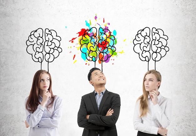 Aziatische zakenman en zijn twee vrouwelijke collega's staan bij een betonnen muur met hersenen erboven getekend. Een van de hersenschetsen is kleurrijk.