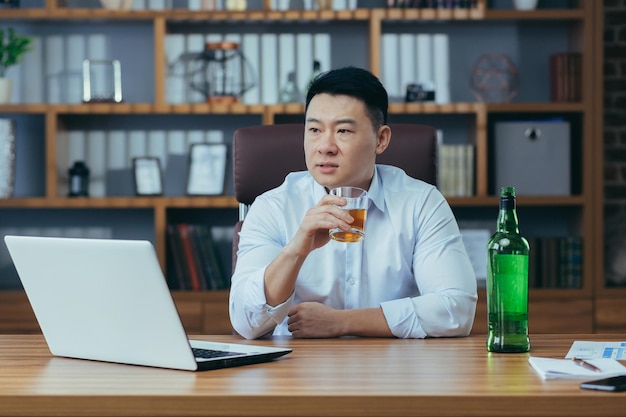 Aziatische zakenman faalt man in wanhoop drinkt sterke drank die laat aan tafel op kantoor zit depressief en hopeloos