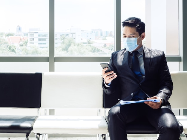 Aziatische zakenman in pak dragen van beschermend gezichtsmasker met behulp van mobiele telefoon zittend in een modern kantoorgebouw in de buurt van enorme glazen raam.