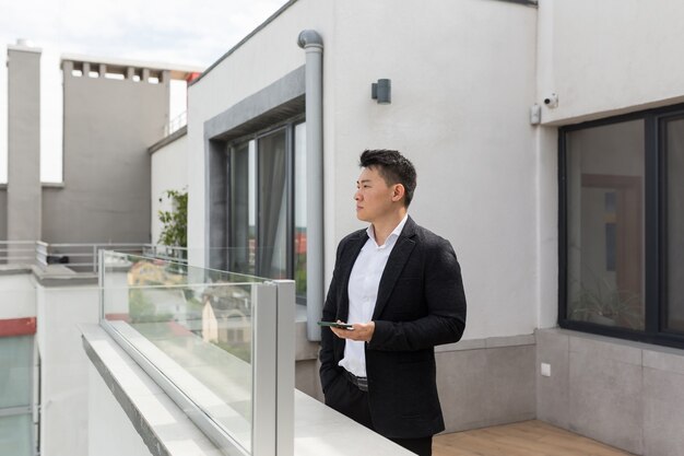 Aziatische zakenman in pak geniet van telefoon op balkonterras van modern kantoorcentrum