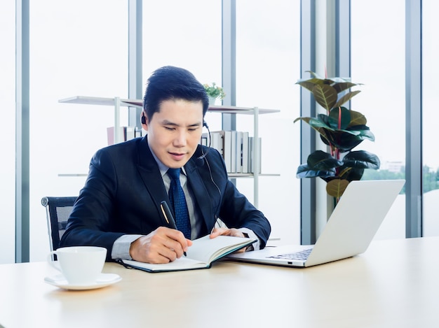 Aziatische zakenman in pak met hoofdtelefoon die laptop op bureau in bureau met behulp van