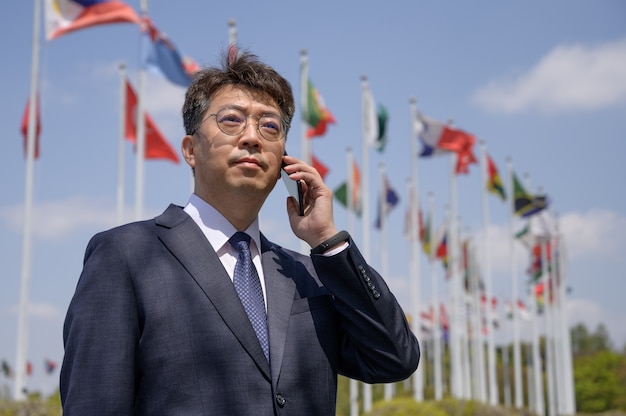 Aziatische zakenman van middelbare leeftijd die een smartphone gebruikt onder verschillende nationale vlaggen die in de wind wapperen.
