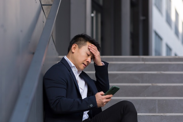 Aziatische zakenmanmakelaar meldt slecht nieuws terwijl hij aan de telefoon praat, in de buurt van kantoor op de trap zit, depressief de hoop verliest en zijn baan failliet gaat