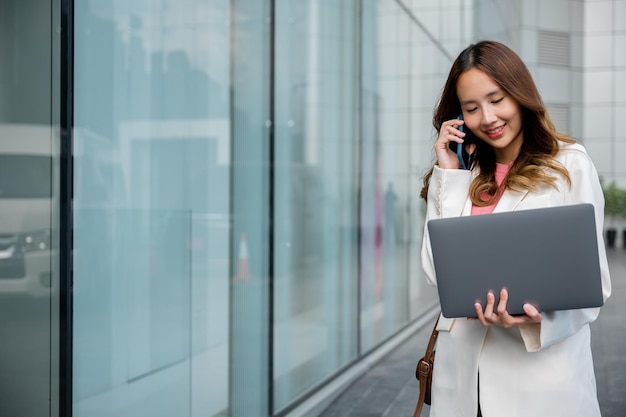 Aziatische zakenvrouw die op laptop werkt en op mobiele telefoon praat aan de voorkant van het gebouw in de buurt van kantoor, Portret mooie zakenvrouw in wit pak glimlach die problemen bespreekt op slimme mobiele telefoon in de stad