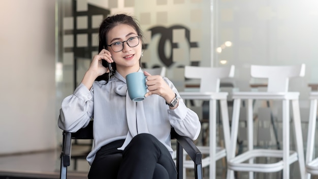 Aziatische zakenvrouw drinken graag koffie en praten aan de telefoon tijdens uw vrije tijd op kantoor.