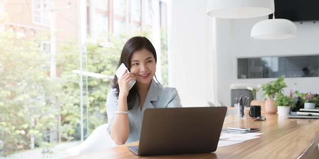 Aziatische zakenvrouw heeft het plezier om te praten op de telefoon, laptop en tablet op het bureau