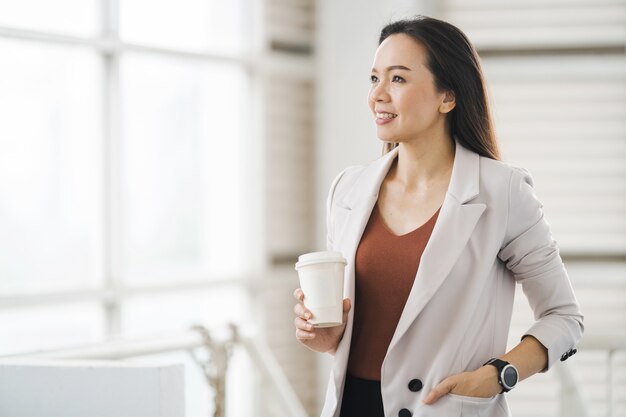 Aziatische zakenvrouw in een ontspannen pak houdt koffiekopjes mee in het bedrijfsgebouw