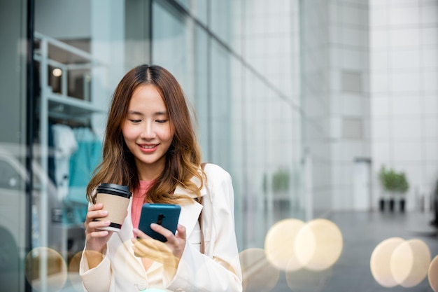 Aziatische zakenvrouw met koffiekopje afhaalmaaltijden kijken smartphone gaan werken lopen op straat in de buurt van kantoor in de ochtend, portret zakenvrouw houden mobiele telefoon en papieren kopje warme drank buiten