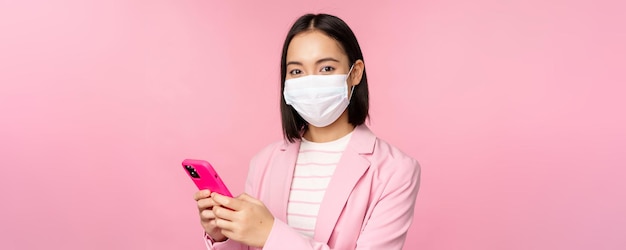 Aziatische zakenvrouw met medisch gezichtsmasker met mobiele telefoon Japenese verkoopster zakelijke dame in pak met smartphone die over roze achtergrond staat