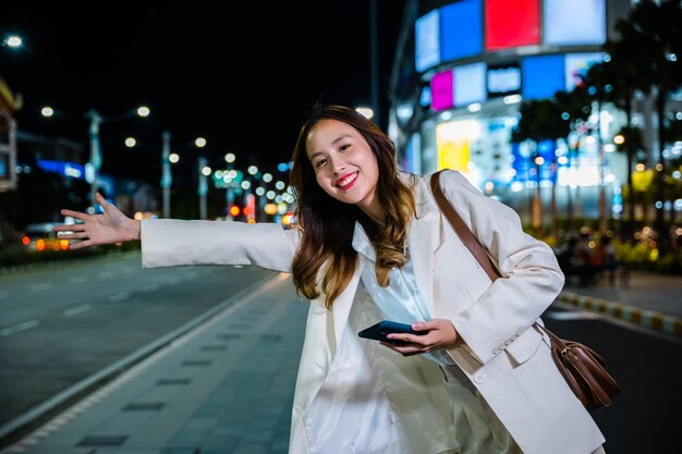 Aziatische zakenvrouw staande hagel zwaaiende handtaxi op weg in drukke stadsstraat 's nachts, mooie vrouw die lacht met behulp van mobiele telefoontoepassing begroetend met hand omhoog bellende taxi na laat werk