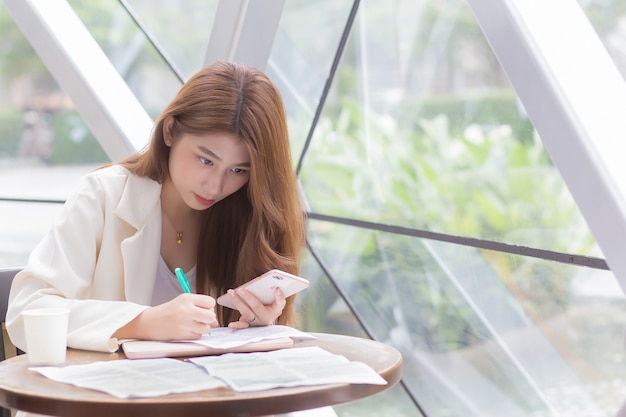 Aziatische zelfverzekerde vrouw draagt een crèmekleurig pak terwijl ze een smartphone gebruikt en iets schrijft