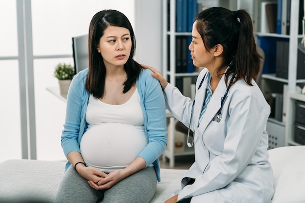 Aziatische zwangere vrouw die aan depressie lijdt, kijkt naar haar psycholoog-arts die op haar schouder klopt en steun toont tijdens consultatie in de kliniek