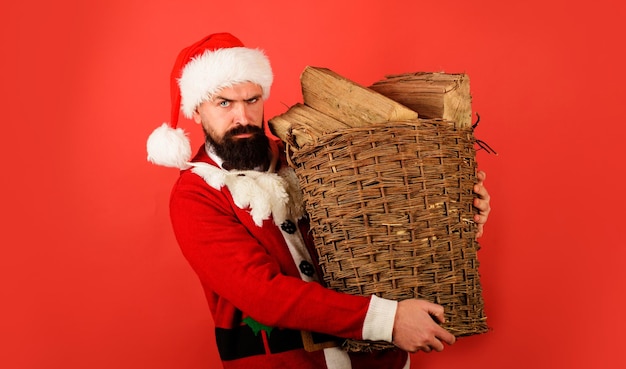 Foto baarde man in een kerstmankostuum die een mand met gehakt brandhout voor de wintervakantie in de open haard draagt