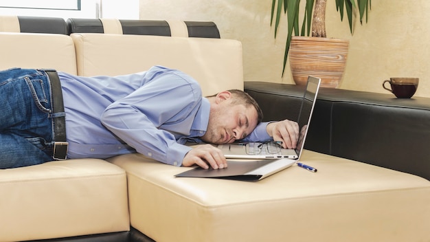 Baas zakenman in een blauw shirt viel in slaap op de bank voor een laptop. Uitgeputte man ontspannen op kantoor. De leidinggevende die de leiding had, viel op het werk in slaap.